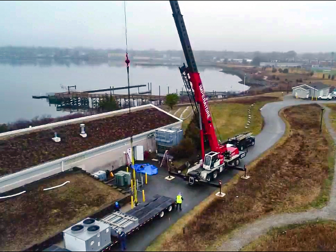 An aerial shot. In the foreground: A red crane parked in front of a long building lifts a large HVAC component off of a flatbed truck along the coast of Fields Point in Providence, R.I. In the background: A foggy pier extends into the waters off Fields Point.