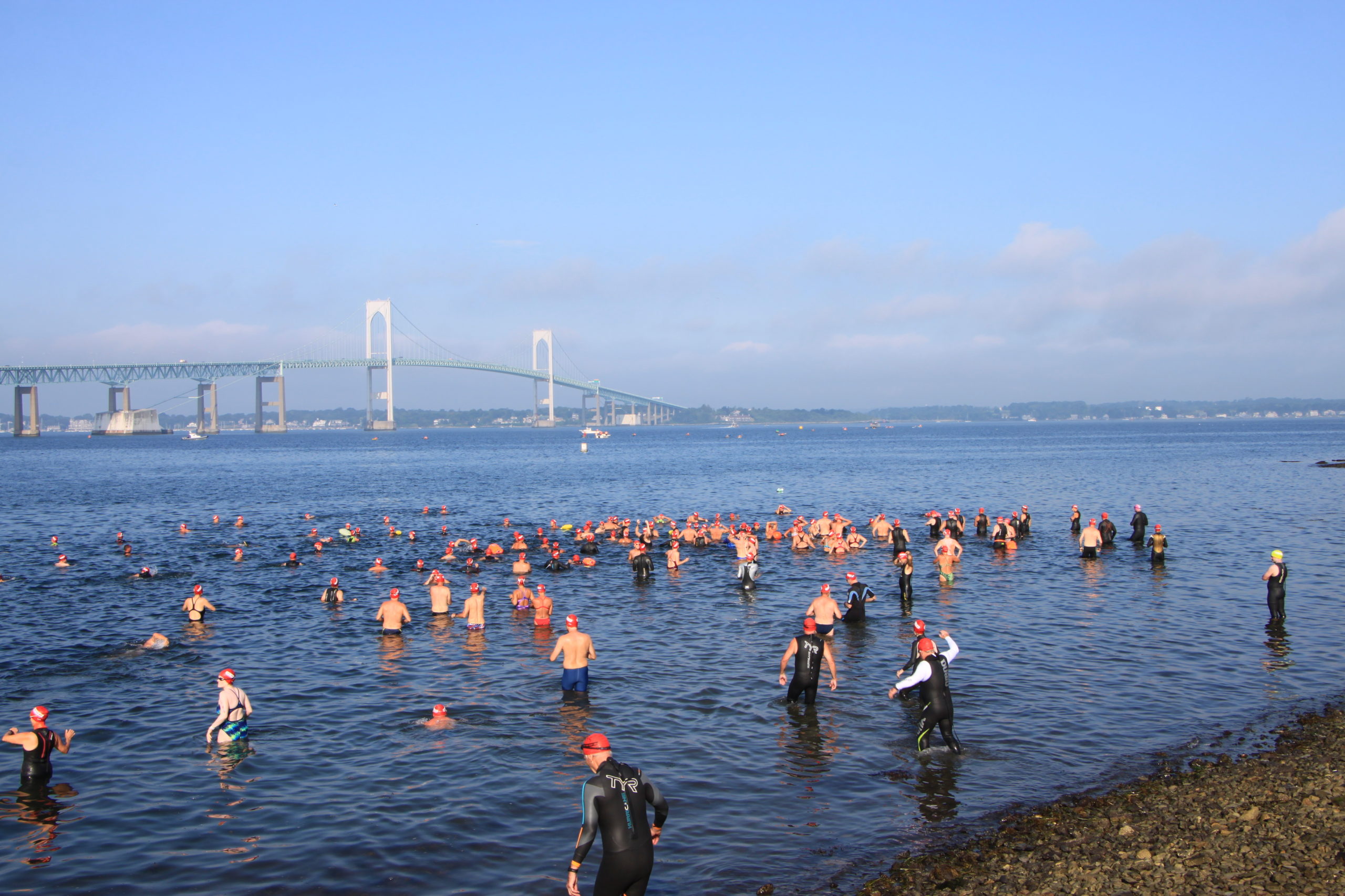 The first wave of swimmers prepares to depart from the Newport shore during the 2022 Save The Bay Swim. The Newport Bridge is visible in the background, as is the Jamestown shoreline.