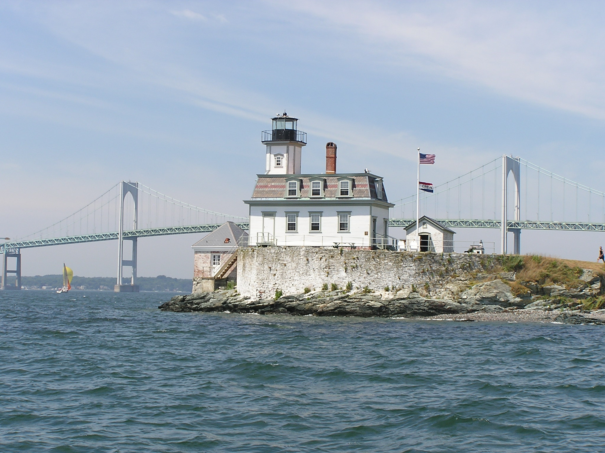 A white lighthouse sits on a stone foundation outcropping on Narragansett Bay. The water reaches the stones at the bottom of the foundation, and the blue-green Newport Bridge is visible in the background